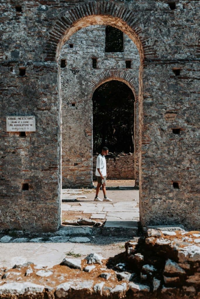 Op een zonnige dag loopt iemand in korte broek en met een hoed op door de stenen boog van oude ruïnes, waarmee hij de sfeer weergeeft van Albanese nomaden die tijdens hun roadtrip door Albanië verborgen pareltjes ontdekken.