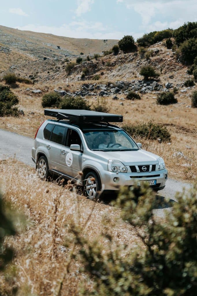 Een zilverkleurige SUV met een dakkoffer rijdt over een smalle landweg door een droog, met gras begroeid landschap met heuvels en hier en daar wat struiken – een klassiek tafereel voor een avontuur met Albanese nomaden, dat de sfeer van een echte roadtrip door Albanië perfect weergeeft.