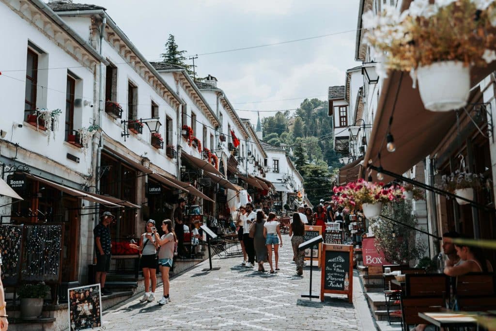 Op een zonnige dag wandelen mensen door een met kinderkopjes geplaveide straat in een historisch Albanees stadje, waar witte gebouwen, cafés en winkels de straat omzomen – een perfecte tussenstop voor iedereen die met Roadtrip Door Albania op ontdekkingstocht gaat of op zoek is naar de charme die Albanees reizigers zo aantrekt.