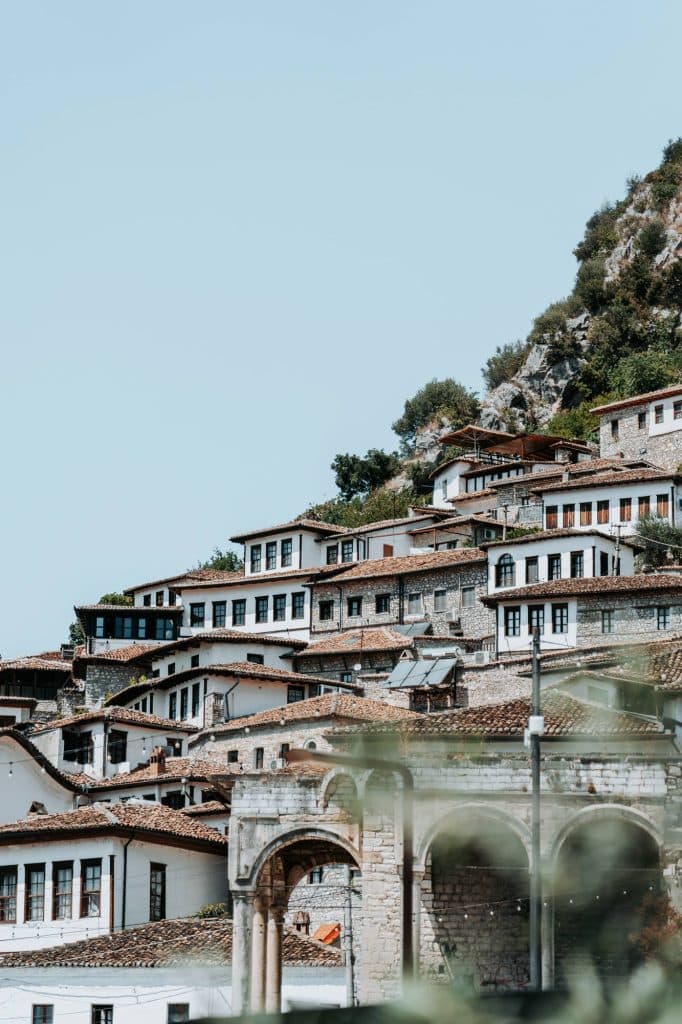 Een groepje traditionele witte stenen huisjes met bruine pannendaken, gebouwd op een heuvel, omgeven door groen onder een heldere hemel – het perfecte decor voor een authentieke roadtrip door Albanië, waar Albanese nomaden al sinds jaar en dag rust vinden te midden van dit tijdloze landschap.