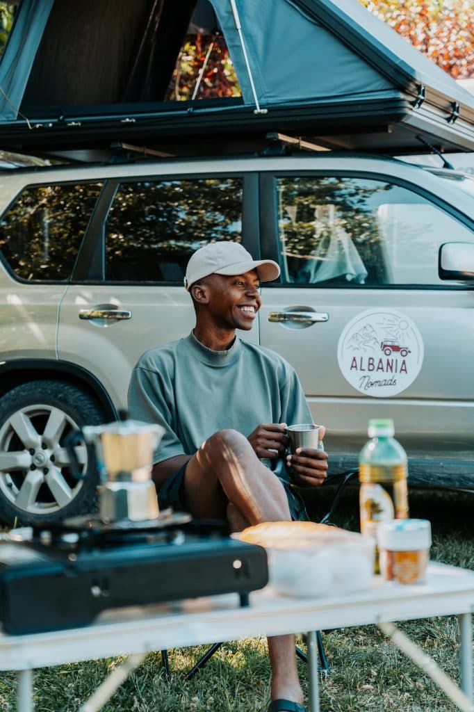 Een persoon zit glimlachend met een mok in de hand voor een auto met een daktent. Hun kampeerspullen en etenswaren liggen uitgestald op een tafel op de voorgrond, wat de avontuurlijke sfeer van een roadtrip door Albanië perfect weergeeft – een authentiek kijkje in het leven van de Albanese nomaden.