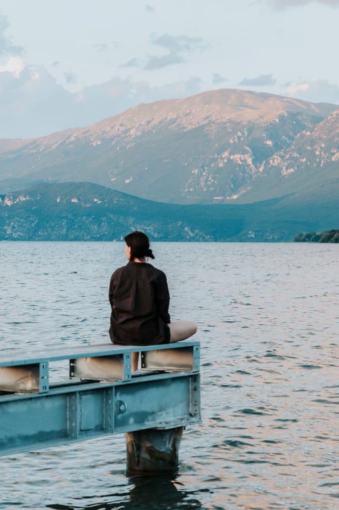 Een persoon zit op de rand van een steiger, met uitzicht op een uitgestrekt wateroppervlak en bergen op de achtergrond, in zacht daglicht — een beeld dat de avontuurlijke geest van de Albanese nomaden tijdens een roadtrip door Albanië weergeeft.