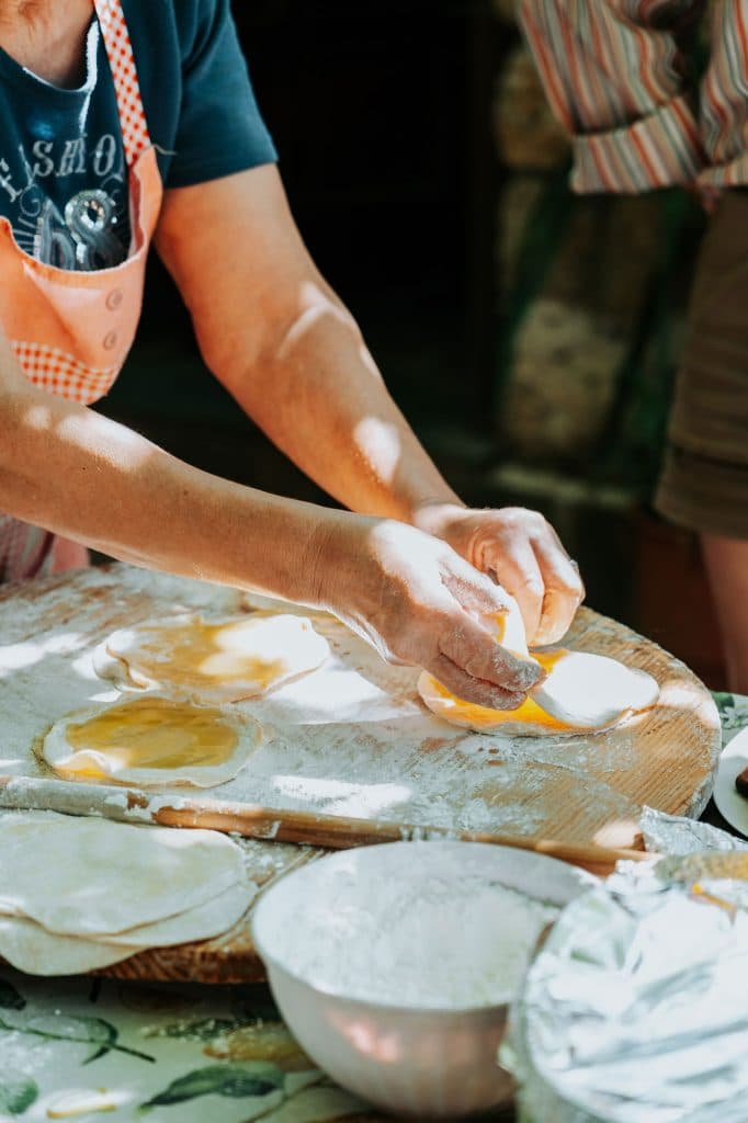 Iemand die op een houten plank deeg bereidt met bloem en ei, en daar cirkels van vormt om te bakken – een tafereel geïnspireerd op de culinaire tradities die je vaak tegenkomt tijdens een roadtrip door Albanië. Op de tafel staan kommen met bloem en andere ingrediënten, die doen denken aan de huiselijke maaltijden waarvan Albanese nomaden tijdens hun reizen genieten.