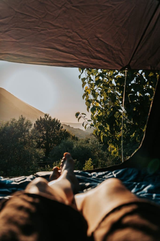 Een persoon die in een tent ligt, met de benen zichtbaar op de voorgrond, terwijl hij uitkijkt op een schilderachtige zonsopgang boven bomen en heuvels – een perfecte ochtend voor Albanese nomaden tijdens een roadtrip door Albanië.