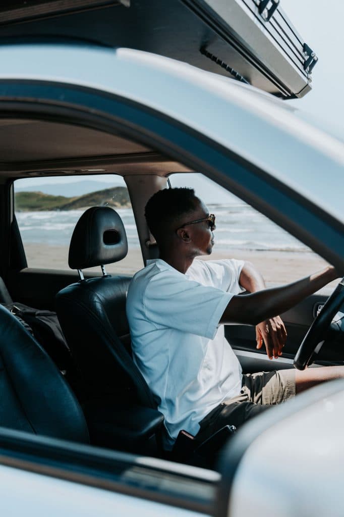 Een persoon met een zonnebril en een wit T-shirt zit op de bestuurdersstoel van een geparkeerde auto en kijkt door het raam naar een strandtafereel – een perfect ‘roadtrip-moment in Albanië’ zoals veel Albanië-nomaden dat zoeken.