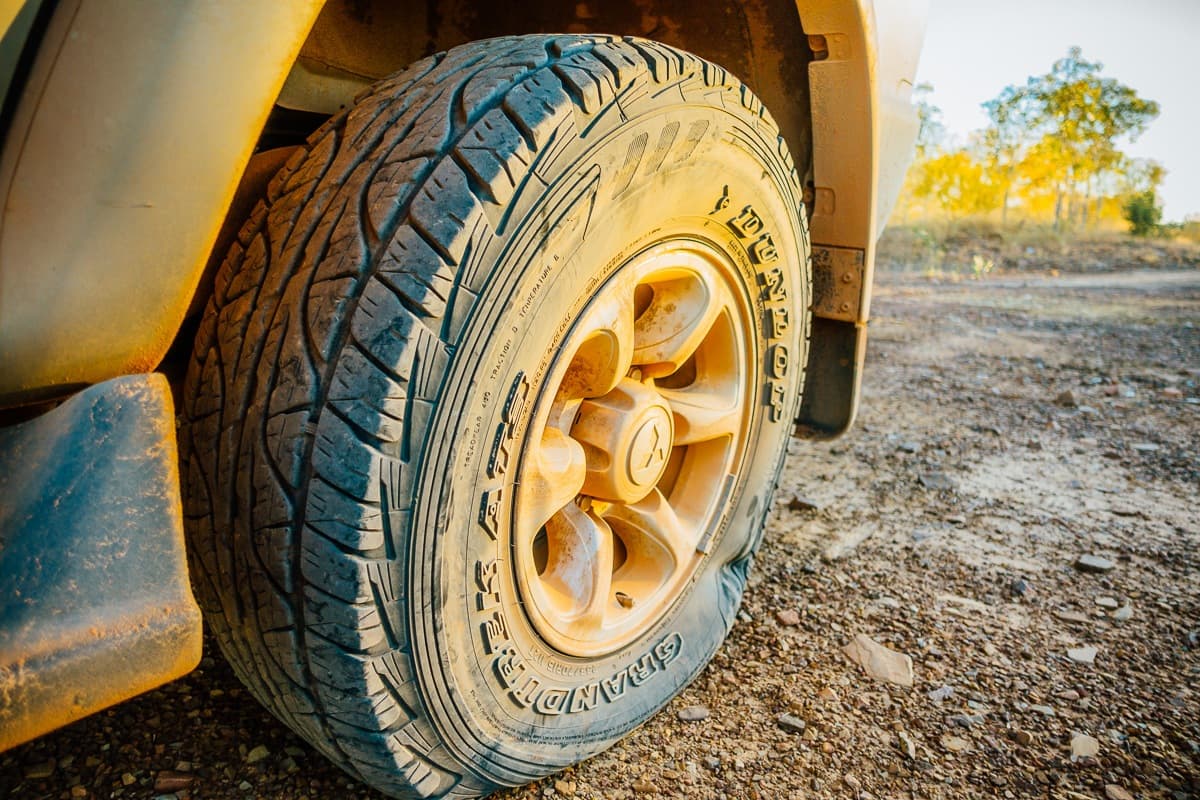 Close-up van een stoffige, ruige band gemonteerd op een voertuig langs de Savannah Way - perfect voor een roadtrip in Australië. Het diepe profiel snijdt door geelbruine modder terwijl het zonlicht de rotsachtige grond en verre bomen op de achtergrond benadrukt.
