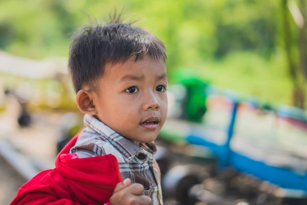Een jonge jongen met kort donker haar, die een bruin geruit overhemd draagt en een rode jas vasthoudt, staat buiten. Hij doet denken aan portretten van Cambodjanen en staart rustig opzij te midden van vaag groen gebladerte en kleurrijke voorwerpen.