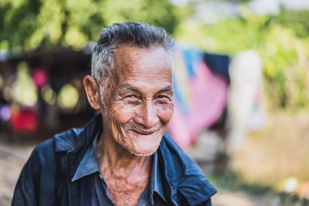 Een oudere man met kort grijs haar glimlacht hartelijk buiten. Hij heeft diepe rimpels, een lichte baard en draagt een donker jasje over een hemd met kraag - een van de portretten van Cambodjanen - omlijst door bomen en kleurrijke doeken in natuurlijk licht.