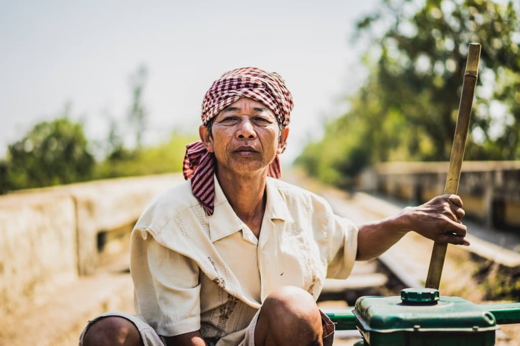 Een oudere persoon met een licht overhemd en een rood-wit geruite sjaal zit buiten en houdt een houten paal vast. De zonovergoten achtergrond met vage bomen doet denken aan portretten van Cambodjanen in een landelijke of agrarische omgeving.