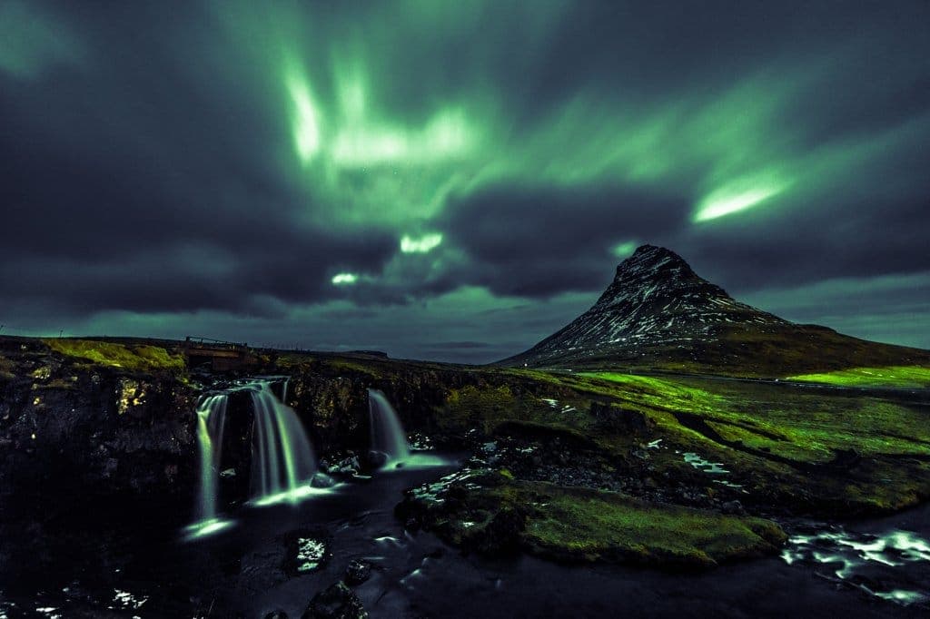 Een dramatisch nachtzicht van de berg Kirkjufell in IJsland in de winter, met daarboven groen noorderlicht. Een kleine waterval stroomt in een donkere rivier, omgeven door bemost, ruig terrein. Het landschap ziet er mysterieus en levendig uit.