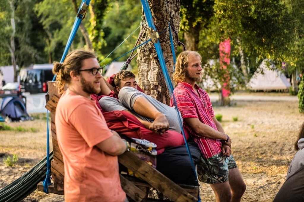Drie mannen relaxen buiten bij bomen op een Surfkamp in Frankrijk. De een zit in een hangmatschommel gewikkeld in dekens, de ander leunt naast hem, terwijl de derde man vlakbij staat. Tenten, een busje en festivalvibes vullen de groene omgeving.