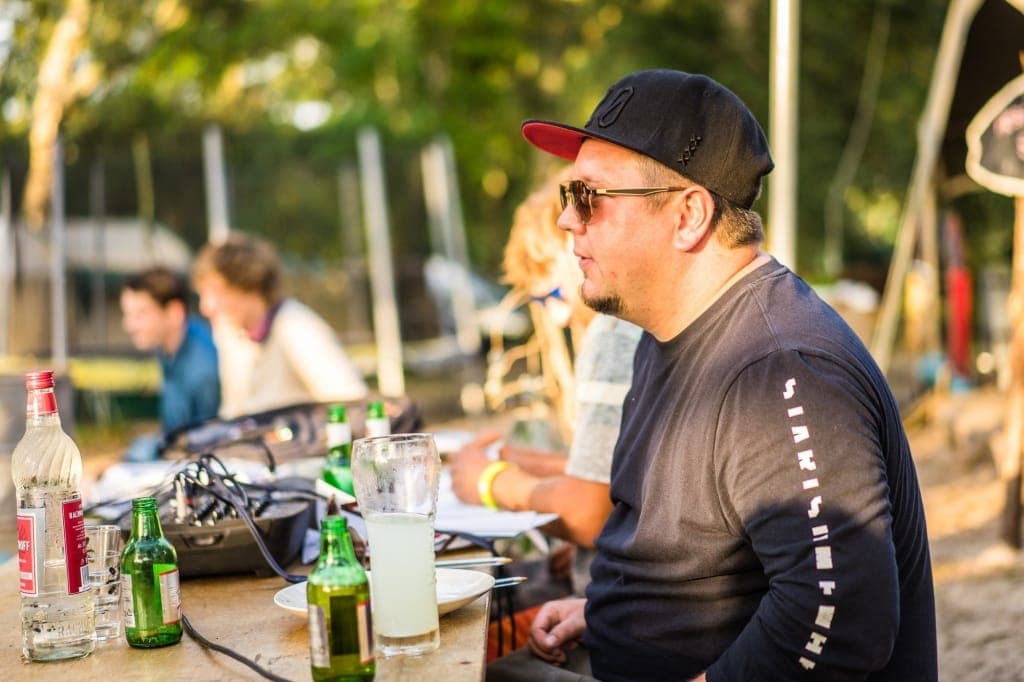 Een man met een zonnebril en een zwarte pet zit met een drankje aan een buitentafel te genieten van een Surfkamp in Frankrijk. Mensen vervagen op de achtergrond onder het warme, gouden zonlicht, waardoor de ontspannen sfeer van dit ongedwongen samenzijn wordt vastgelegd.