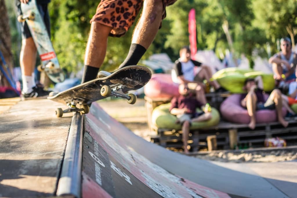 Een close-up van een skateboarder in luipaardprint shorts die een truc uitvoert op een schans tijdens een Surfkamp in Frankrijk, met toeschouwers die ontspannen op kleurrijke zitzakken te midden van zonnige luchten, bomen en groen.