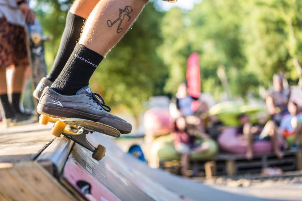Close-up van de benen van een skateboarder, grijze schoenen en zwarte sokken, balancerend op een skateboard aan de rand van een schans. Op de kuit is een tatoeage van een stokfiguur te zien, die de relaxte sfeer van het Surfkamp in Frankrijk oproept tussen vage bomen en zittende mensen.