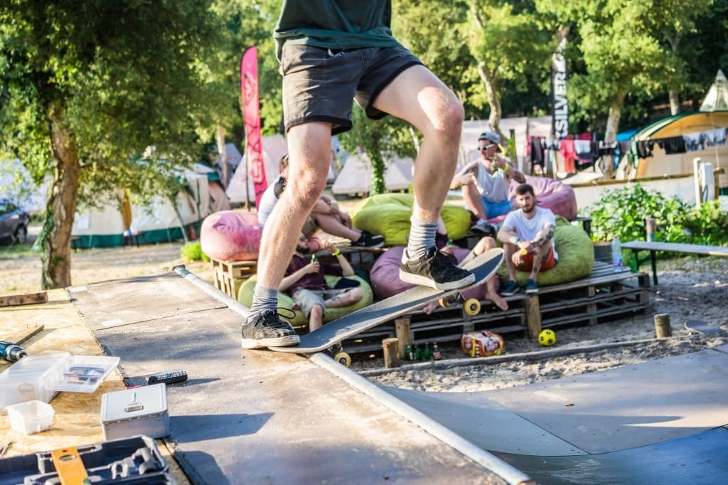 Een skateboarder balanceert buiten op een rail, knieën gebogen, midden in een trick. Vlakbij zitten vier mensen op kleurrijke zitzakken op houten pallets op een Surfkamp in Frankrijk, omringd door bomen, kampeertenten en spullen in de ontspannen, zonnige parksfeer.