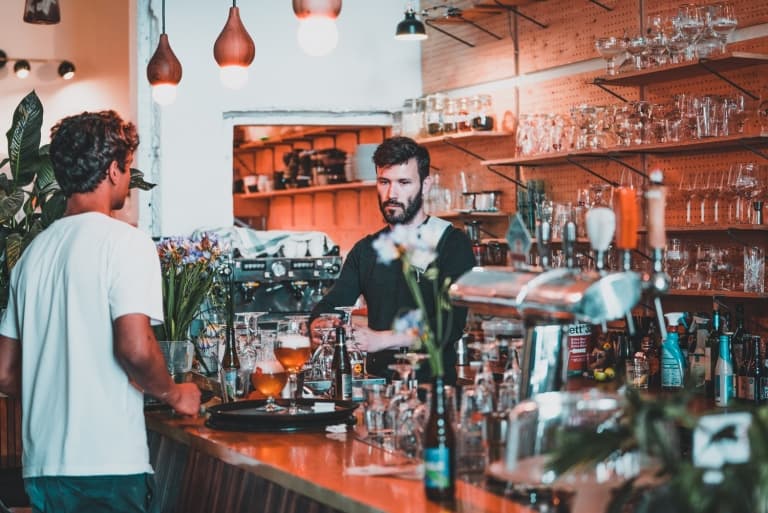 Een bebaarde barman staat achter een houten bar tegenover een mannelijke klant in een wit overhemd. Glazen en flessen staan op de planken, met hanglampen erboven. De ongedwongen, moderne sfeer weerspiegelt het relaxte gevoel van slapen op een laars in Mechelen.