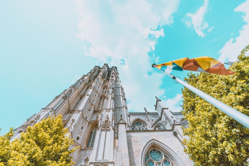 Een lage hoek van een grote gotische kathedraal met sierlijk steenwerk, hoge boogramen en een toren. Bij groene bomen onder een heldere hemel wappert een vlag - een sereen gezicht na het slapen op een laars in Mechelen.