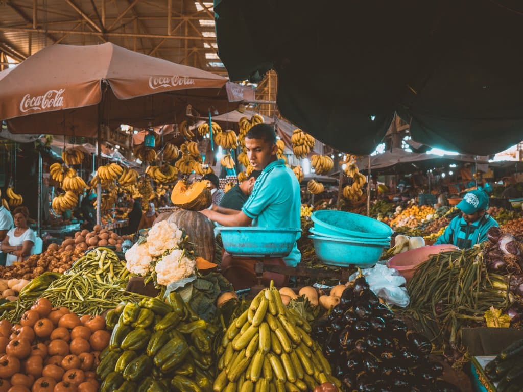 Een man in een blauw shirt snijdt pompoen op een drukke, kleurrijke markt geïnspireerd op de souks van Marokko. Hij wordt omringd door fruit en groenten - bananen, papaja's, aubergines - met blauwe schalen in de buurt en marktparaplu's boven zijn hoofd, terwijl verkopers en winkelend publiek druk in de weer zijn.