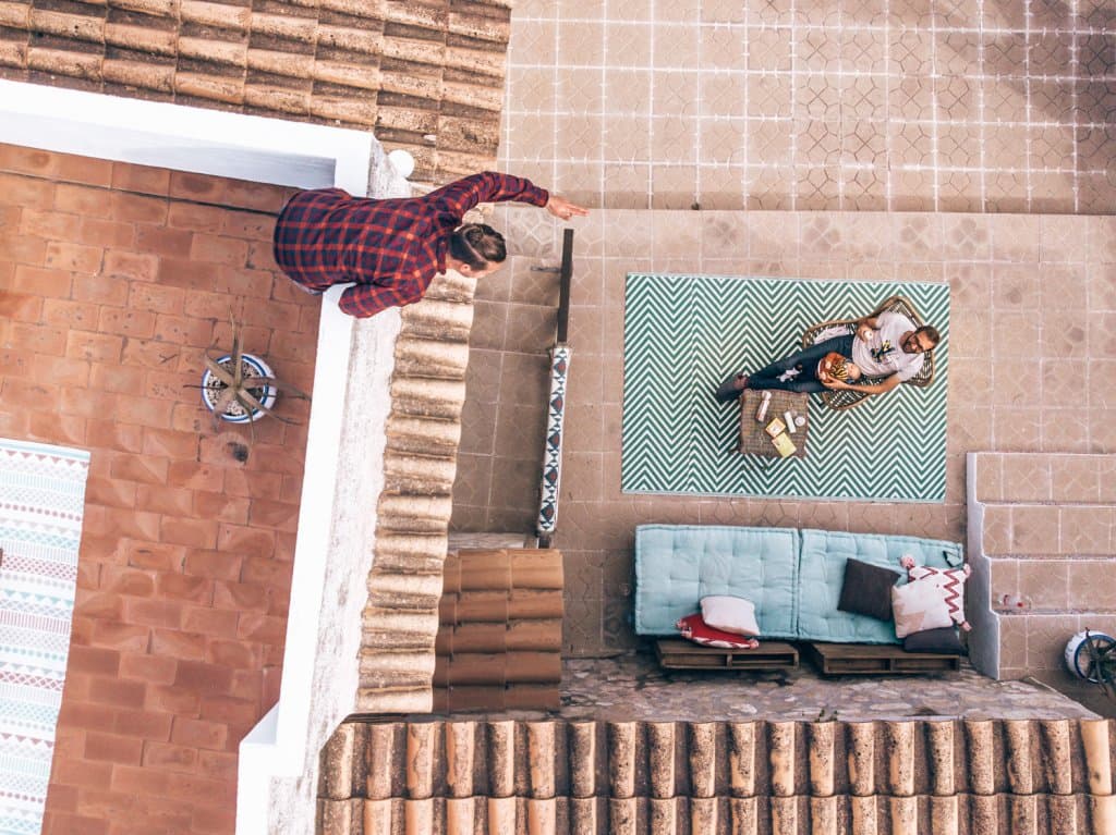 Luchtfoto van twee mensen die ontspannen op een groen gedessineerd tapijt naast een blauwe bank in een ecoresort in Spanje. Een andere persoon in een rood geruit overhemd leunt over een richel op het dak boven hen, met buitenmeubilair en tegels met patronen die de scène compleet maken.
