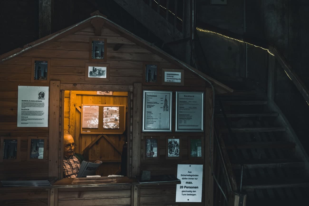 Een man zit in een klein houten kaartjeshokje in Goslar en leest een krant. Het warm verlichte hokje is versierd met posters en foto's, terwijl een bord in het Duits over kaartverkoop mensen verwelkomt die deze charmante plek willen bezoeken. Daarachter doemt een donkere trap op.