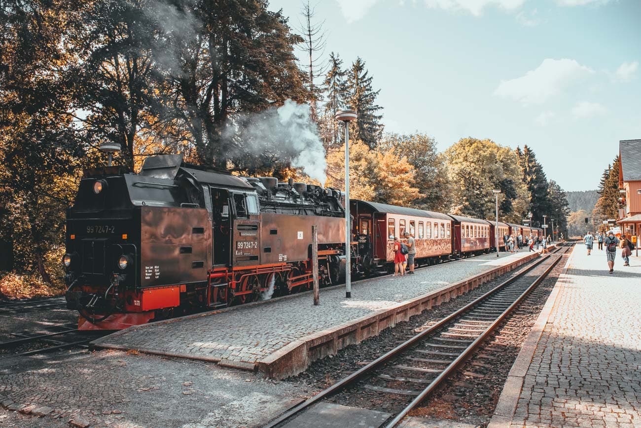 Een oude zwarte stoomlocomotief met rook uit de schoorsteen staat op het perron van een geplaveid treinstation in Goslar; bomen met herfstbladeren omringen het tafereel terwijl mensen langs het perron genieten van hun bezoek onder een heldere hemel.