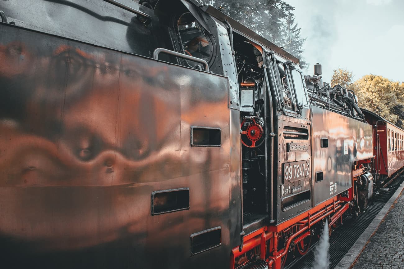 Close-up van een zwart-rode stoomlocomotief op een treinstation bij Goslar bezoeken, met gedetailleerd metaalwerk en zichtbare machines. Stoom drijft omhoog terwijl bomen en een deel van het perron op de achtergrond te zien zijn.