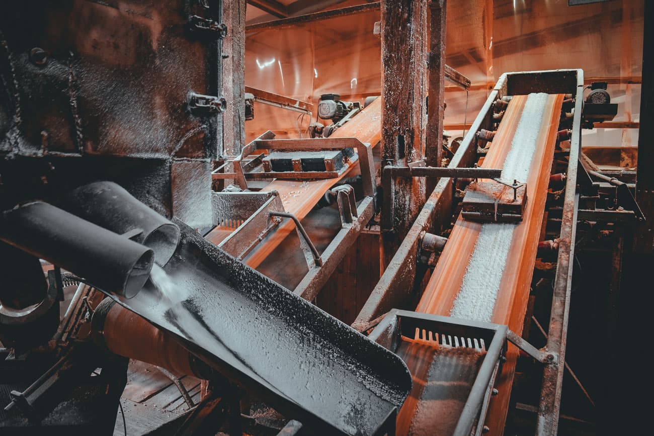 Industriële scène met meerdere transportbanden die een wit poederachtig materiaal transporteren in een fabriek. De machines zijn metaalachtig en versleten, met oranje verlichting die een warme gloed werpt. Het doet denken aan locaties die je misschien ziet als je goslar bezoekt.