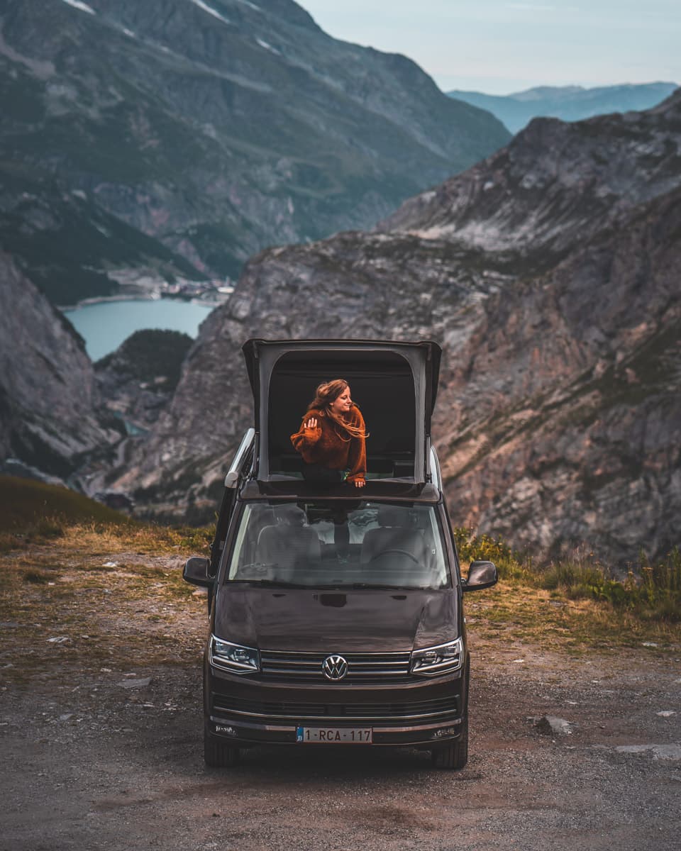Een persoon in een oranje jas staat in het open dak van een zwart Volkswagenbusje tijdens een roadtrip door de Franse Alpen. Majestueuze rotsachtige bergen en een kronkelend meer of rivier strekken zich uit onder een bewolkte hemel op de achtergrond.