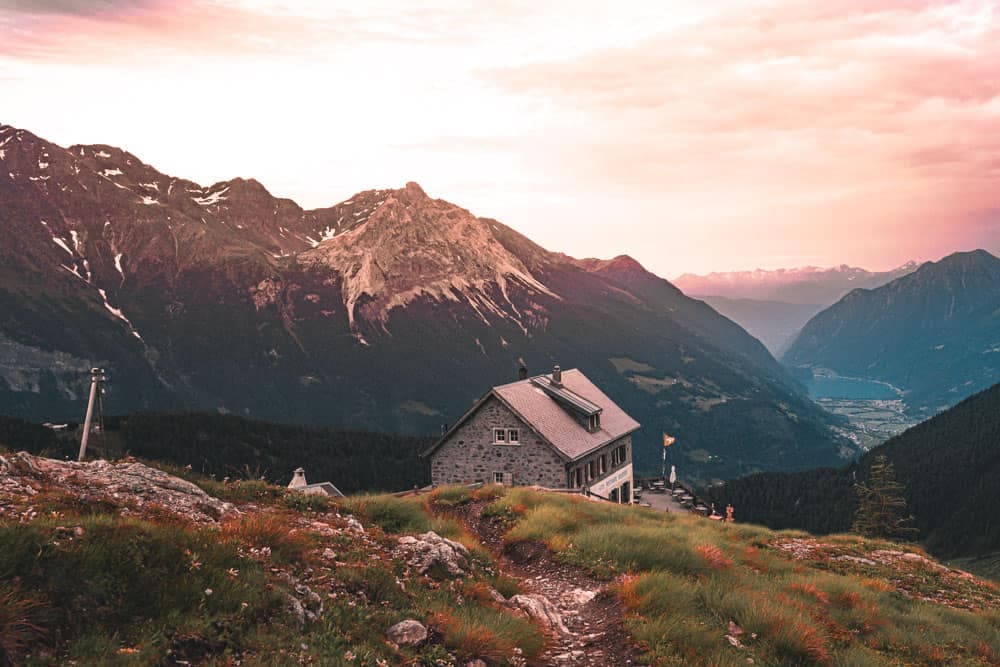 Een stenen berghut met een donker dak staat op een grashelling bij zonsondergang, omringd door wilde bloemen. Majestueuze bergen rijzen erachter op onder een roze en oranje hemel - een idyllisch tafereel om te ontdekken in Zwitserland met de trein.