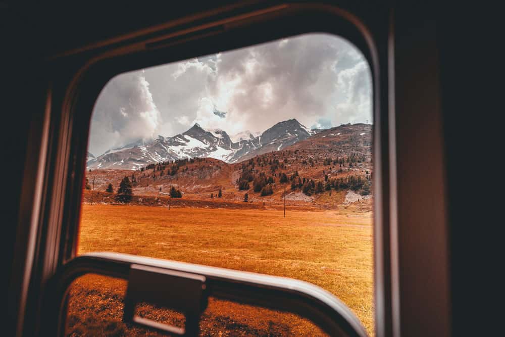 Zicht door een treinraam op een schilderachtig landschap met grasvelden, verspreide bomen en besneeuwde bergen onder een bewolkte hemel - perfect voor reizen door Zwitserland met de trein. Het raamkozijn en een deel van het interieur van de trein zijn zichtbaar op de voorgrond.