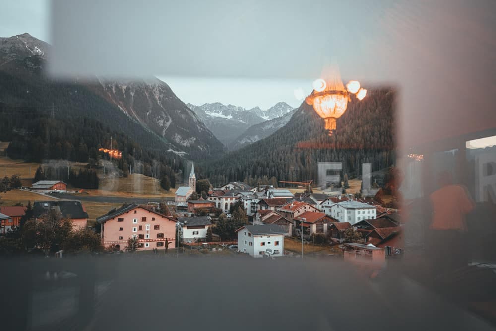 Een bergdorp met verspreide huizen en een kerk wordt gezien door een reflecterend raam, alsof je op reis bent door zwitserland met de trein, met daarachter bergen en bossen. De weerspiegeling van het raam onthult een sierlijke kroonluchter, waardoor de elegantie van het interieur samensmelt met het prachtige uitzicht buiten.
