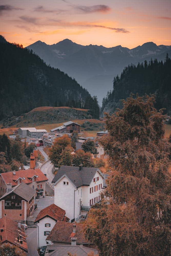 Een schilderachtig bergdorpje bij zonsondergang, met een oranje-roze lucht die de grillige, donkere bergen op de achtergrond verlicht. Gezellige huizen met roodachtige daken nestelen zich tussen de bomen, terwijl kronkelende weggetjes en de charme van zwitserland met de trein uitnodigen tot een rustige verkenningstocht.