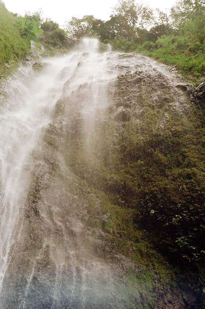 Nevel van een hoge, dunne waterval stroomt naar beneden langs een steile, rotsachtige klif bedekt met groen mos en vegetatie - als een eigen poëzie van de natuur - terwijl bomen en een bewolkte Nicaraguaanse lucht aan de bovenrand van het beeld verschijnen.
