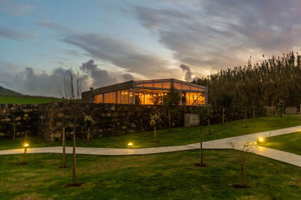 Een modern gebouw met glazen wanden - een van de beste hotels die Sao Miguel te bieden heeft - fonkelt in de schemering met warme binnenverlichting. Een kronkelend geplaveid pad, omzoomd met kleine lichtjes en jonge bomen, leidt door goed onderhouden gras naar het uitnodigende tafereel.