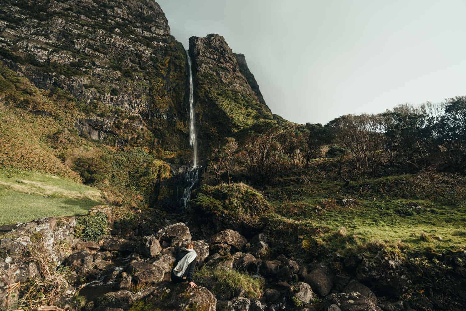 Een persoon in een lichte jas zit op een rotsachtige ondergrond voor een hoge, dunne waterval die langs een weelderige groene berghelling naar beneden stort op Flores, Azoren, omringd door vegetatie en bomen onder een bewolkte hemel - perfect voor liefhebbers van langzaam reizen.