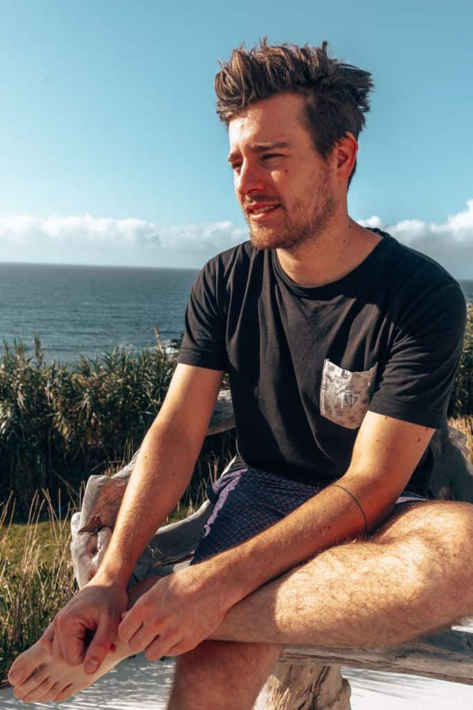 Een jonge man met warrig haar zit buiten op een houten bankje, met zijn benen gekruist en blootsvoets in een zwart T-shirt en korte broek, loensend in het zonlicht - met op de achtergrond de oceaan, planten, blauwe lucht en enkele van de beste hotels die Sao Miguel te bieden heeft.