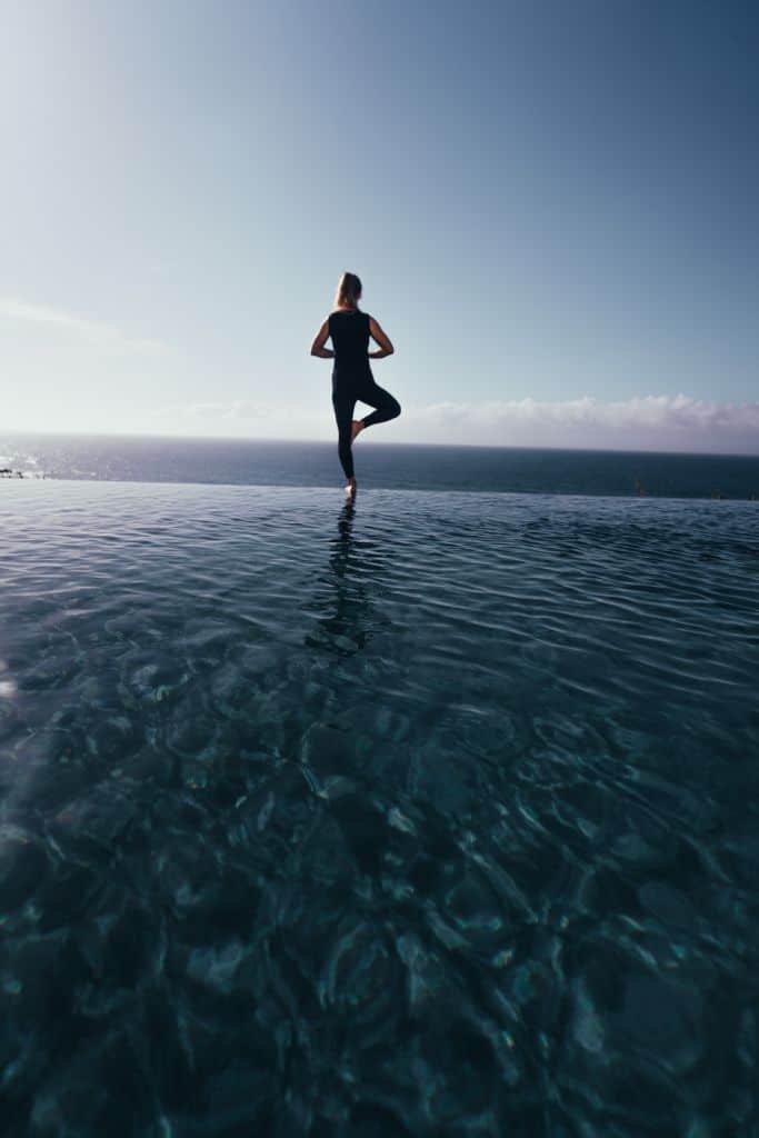 Een persoon in donkere sportkleding staat in boomhouding aan de rand van een overloopzwembad in een van de beste hotels van Sao Miguel, met uitzicht op de oceaan. Het kalme, heldere water weerspiegelt zowel de persoon als de brede blauwe lucht en creëert een vredig en sereen tafereel.