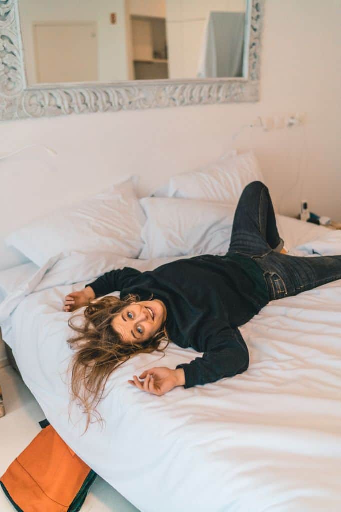 Een vrouw met lang haar, in een donkere trui en spijkerbroek, ligt speels op een wit bed in een lichte, minimale kamer in een van de beste hotels die Sao Miguel te bieden heeft. Ze glimlacht naar een grote decoratieve spiegel; een oranje tas rust op de vloer.