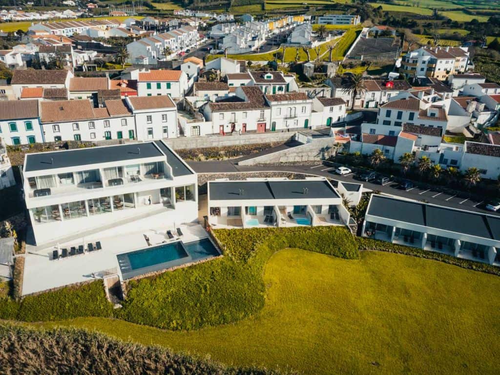 Luchtfoto van een modern wit hotel met een zwembad, terrasstoelen en drie kleinere eenheden op een grashelling. Daarachter staan traditionele huizen met rode daken, terwijl groene velden zich uitstrekken - een van de beste hotels die Sao Miguel te bieden heeft.