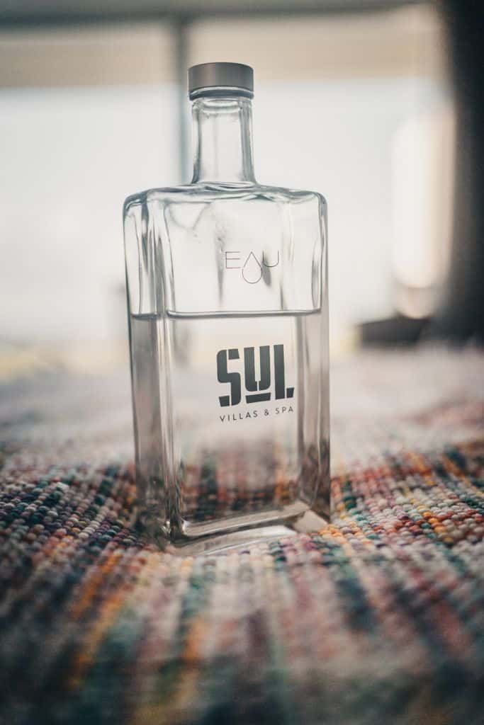 Een heldere, rechthoekige glazen fles half gevuld met water staat op een kleurrijk, geweven oppervlak in een van de beste hotels van Sao Miguel. Op de fles staat "EAU" bij de hals en "SUL VILLAS & SPA" in vette letters. Zacht, natuurlijk licht schijnt op de achtergrond.
