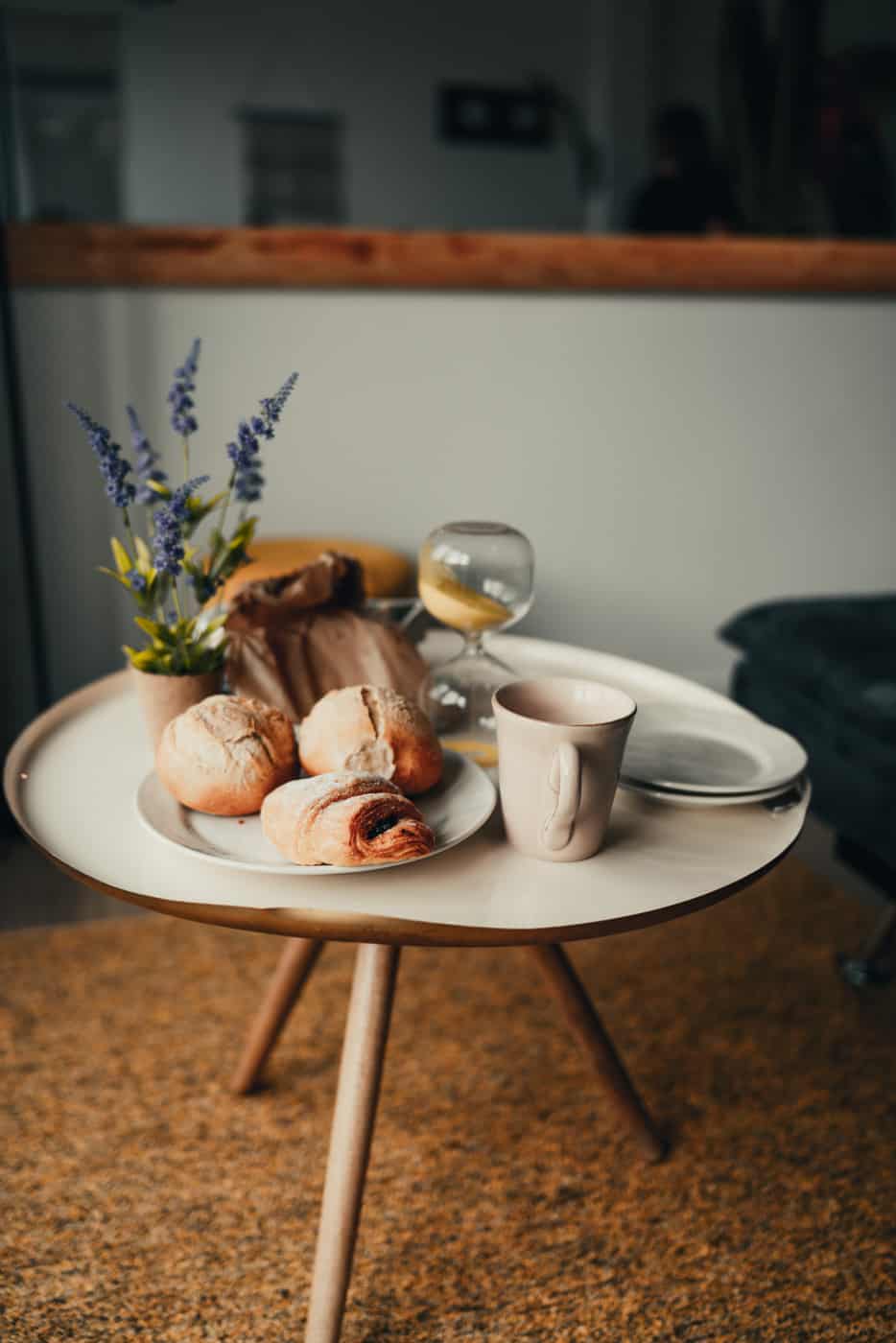 Op een ronde houten tafel, zoals die in de beste hotels op Sao Miguel, liggen broodjes, een gebakje, een witte mok, water met citroen, paarse bloemen in een vaas en bestek op een leeg bord op een taankleurig tapijt in een zacht verlichte kamer.