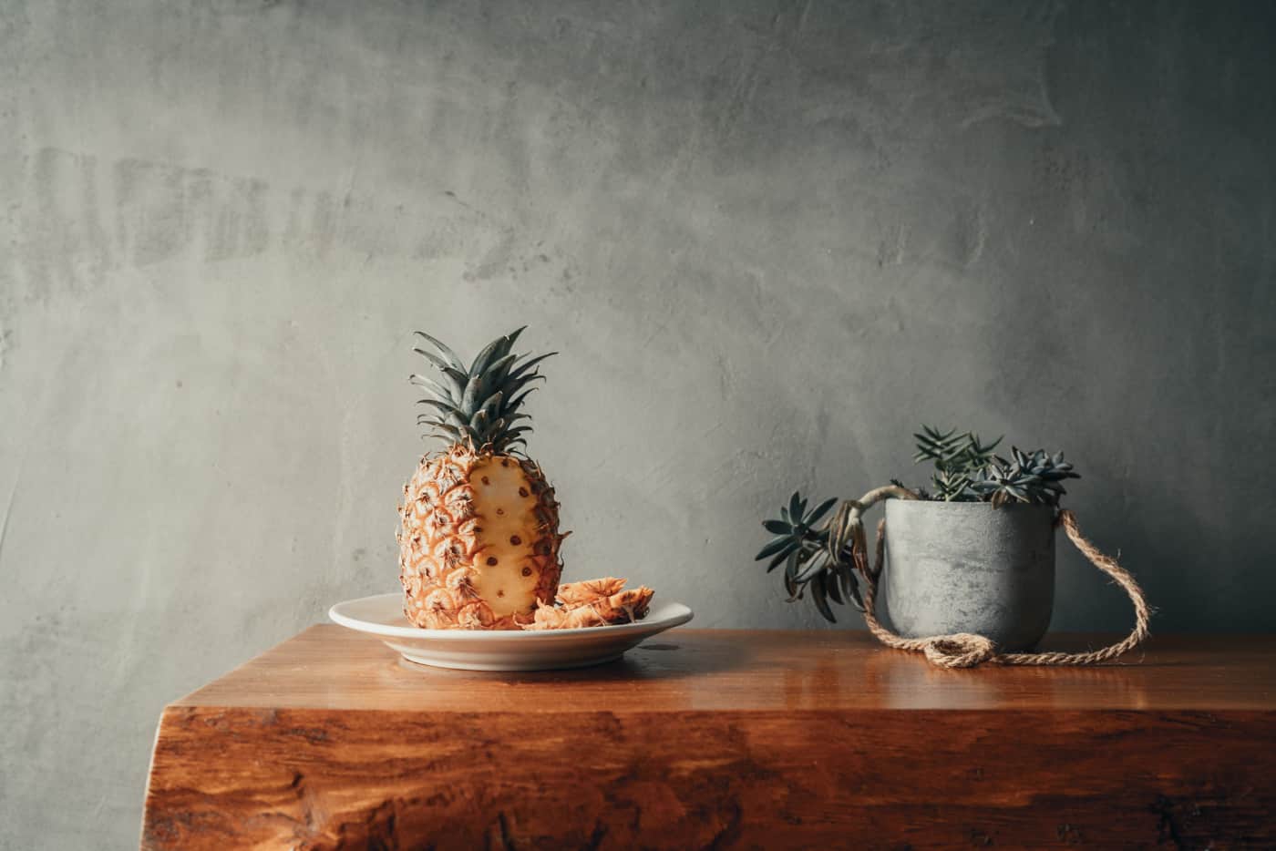 Een rijpe ananas, gehalveerd en in plakjes gesneden, ligt op een wit bord op een houten tafel - perfect om van te genieten na het verkennen van de beste hotels die Sao Miguel te bieden heeft. Rechts staat een kleine vetplant in een grijze pot bij een grijze muur met structuur.