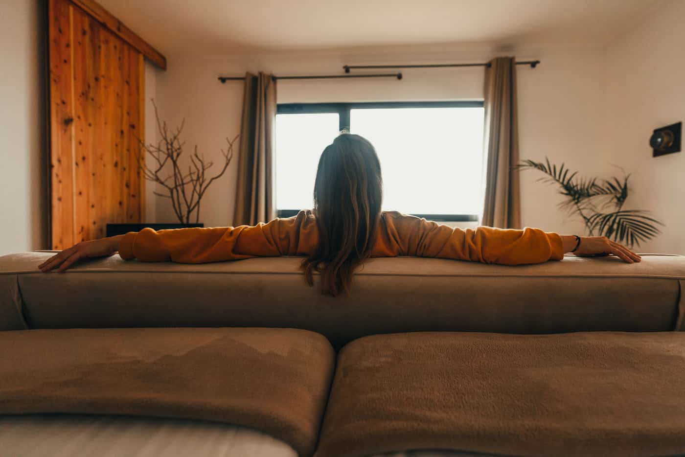 Een persoon met lang haar zit op een beige bank, de armen uitgestrekt langs de rugleuning, met uitzicht op een helder raam met gordijnen - ontspannen alsof ze terugkomen van het verkennen van de beste hotels die Sao Miguel te bieden heeft. De kamer heeft zacht licht en een natuurlijke inrichting.