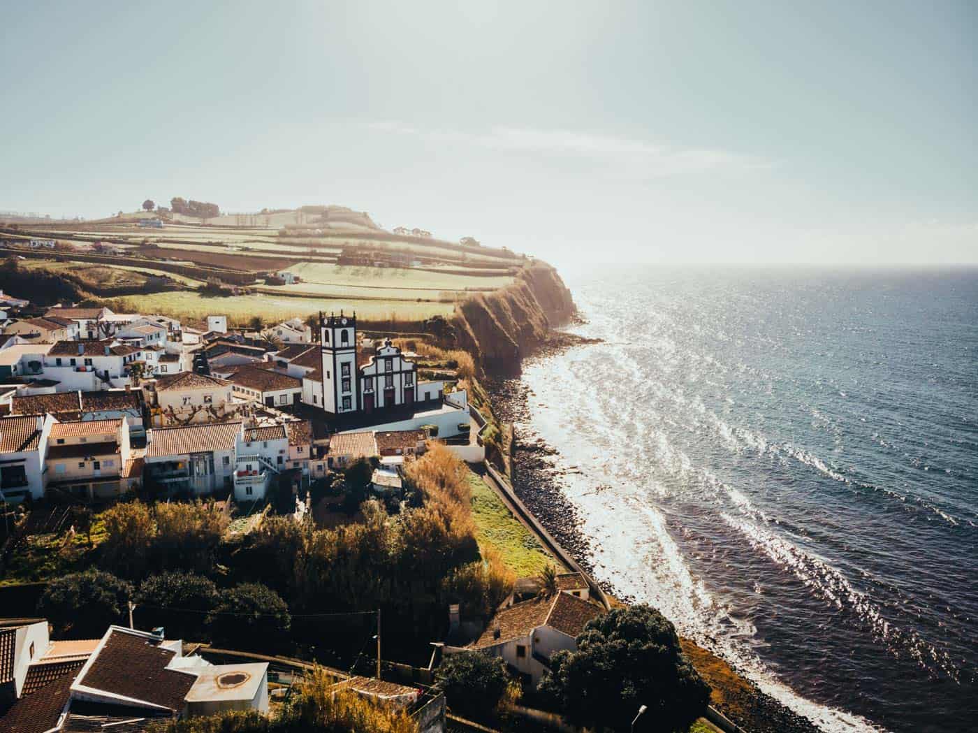 Een kustdorpje met witte huizen en een zwart-witte kerk ligt op een klif met uitzicht op de oceaan, vlakbij enkele van de beste hotels die Sao Miguel te bieden heeft. Golven slaan zachtjes tegen de rotsachtige kust terwijl groene velden zich uitstrekken onder een heldere, heldere hemel.
