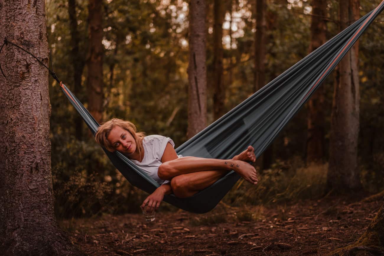 Een vrouw met licht haar en een wit overhemd ligt ontspannen in een donkergroene hangmat tussen de bomen in het Ardennenbos. Glimlachend, de benen opgerold en het glas in de hand, geniet ze van een vredige pitstop terwijl warm gouden licht door de bomen filtert.