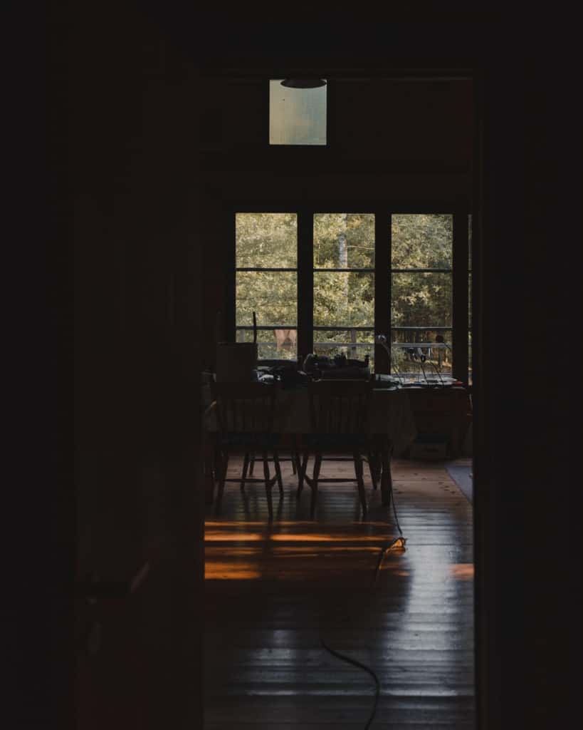 Zonlicht stroomt door hoge ramen op een houten vloer in La Cabane, een gezellige pitstop in de Ardennen. Vanuit de deuropening nodigt een tafel met verspreide voorwerpen en stoelen je uit om binnen te komen, terwijl bomen buiten de zacht verlichte kamer lonken.