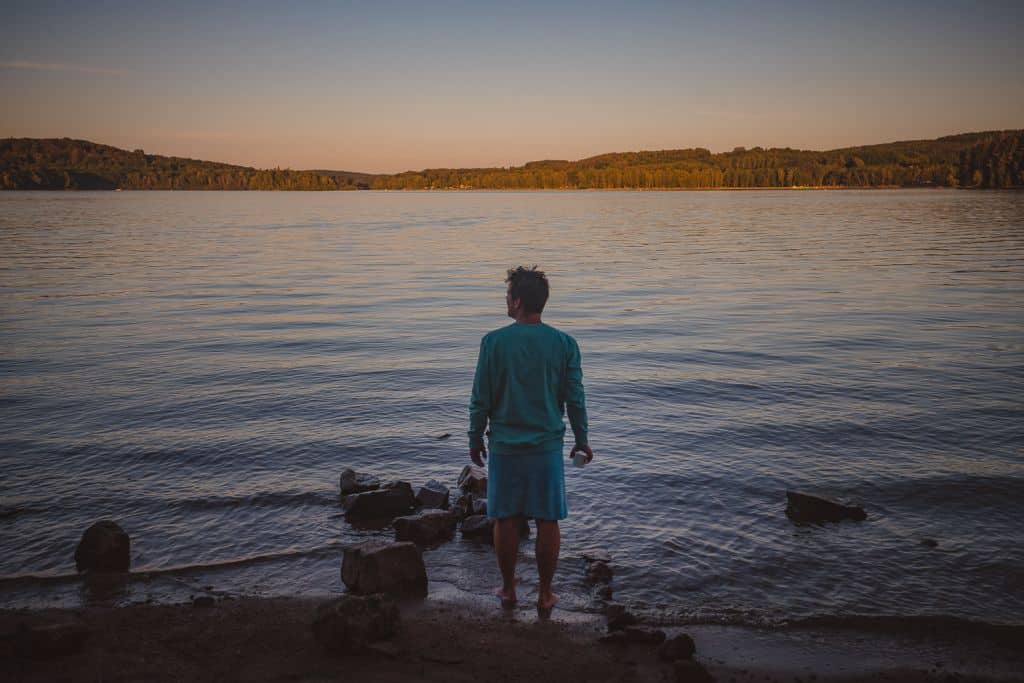 Een persoon met kort donker haar, een groenblauw shirt met lange mouwen en een lichtblauwe korte broek, staat blootsvoets bij een rustig meer in Morvan en kijkt uit over het water. Het gouden avondlicht schijnt over de meren en beboste oevers in de verte.