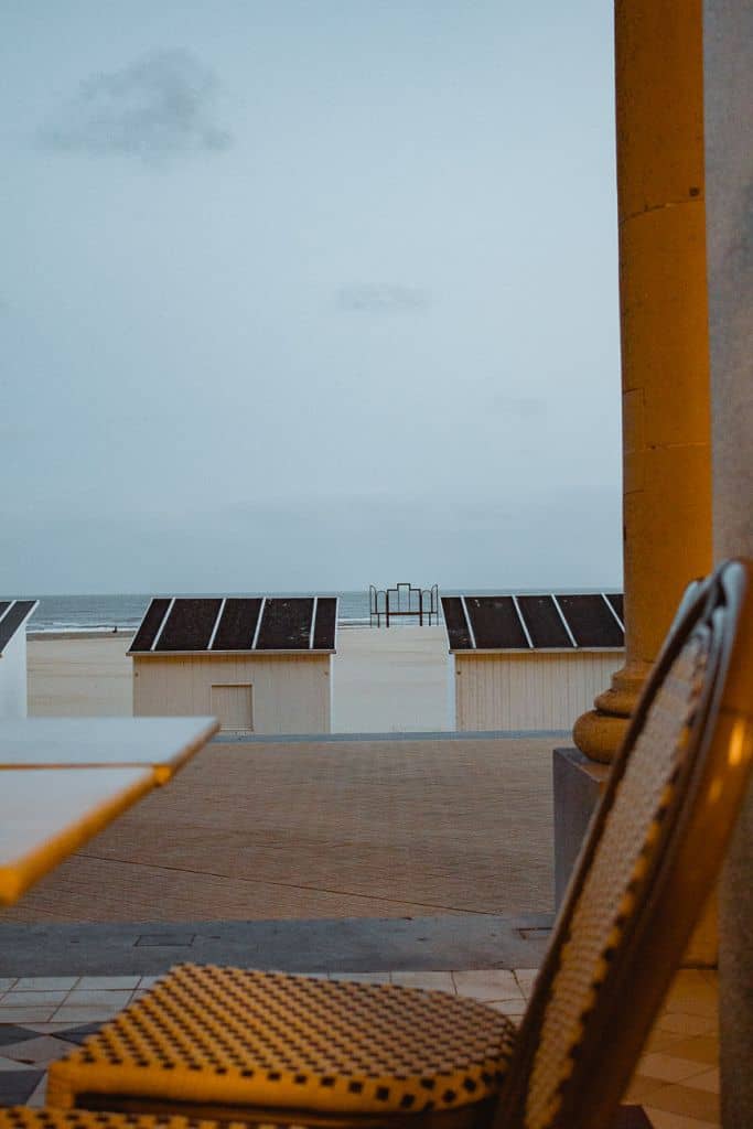 Een stoel met patroon en een tafel staan op een tegelvloer in Oostende, met een zandstrand, kleine witte strandhuisjes en de oceaan daarachter onder een bewolkte hemel in de schemering - een uitnodigend tafereel voor een ontmoeting met een oude vriend. Rechts staat een stenen pilaar.