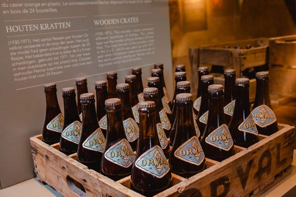 Een houten krat bevat 24 bruine Orval bierflessen met blauwe en oranje etiketten. Achter de krat staat een informatiedisplay in het Nederlands en Engels over houten kratten, die een gevoel van Ardense overlevingsgeschiedenis oproept; een andere krat staat op de achtergrond.