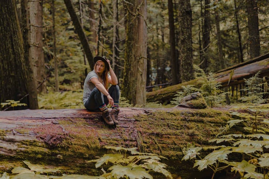 Een vrouw met een hoed op, wandelschoenen aan en kleurrijke sokken aan zit op een grote omgevallen bemoste boomstam in een weelderig bos in Banff National Park, omringd door varens en hoge bomen. Ze kijkt naar boven met een ontspannen uitdrukking en laat één hand op haar hoed rusten.
